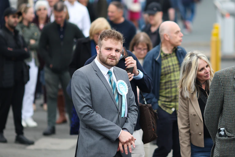A man wearing a Reform rosette standing in a queue.