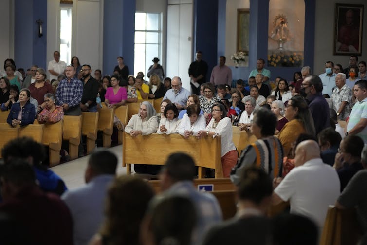 People kneel at pews made of light-colored wood inside a room with blue and white walls.