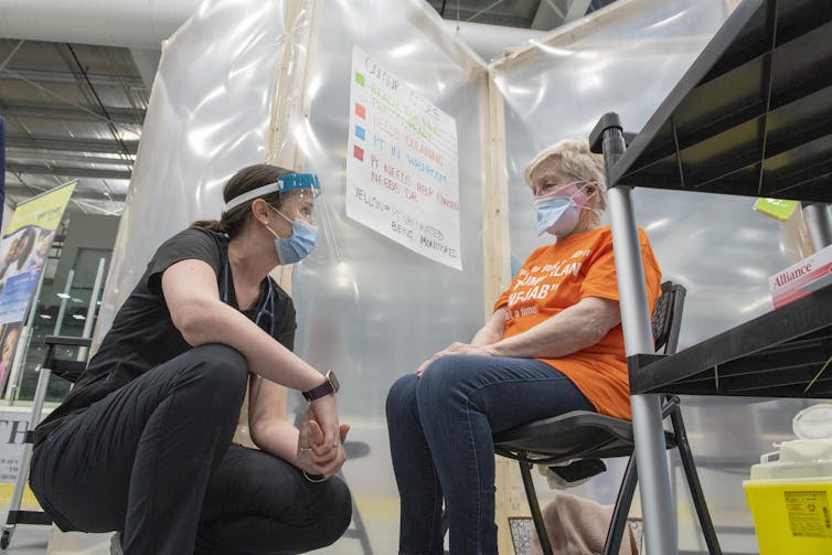 A crouching woman speaking to another seated on a chair. Both wear face masks.