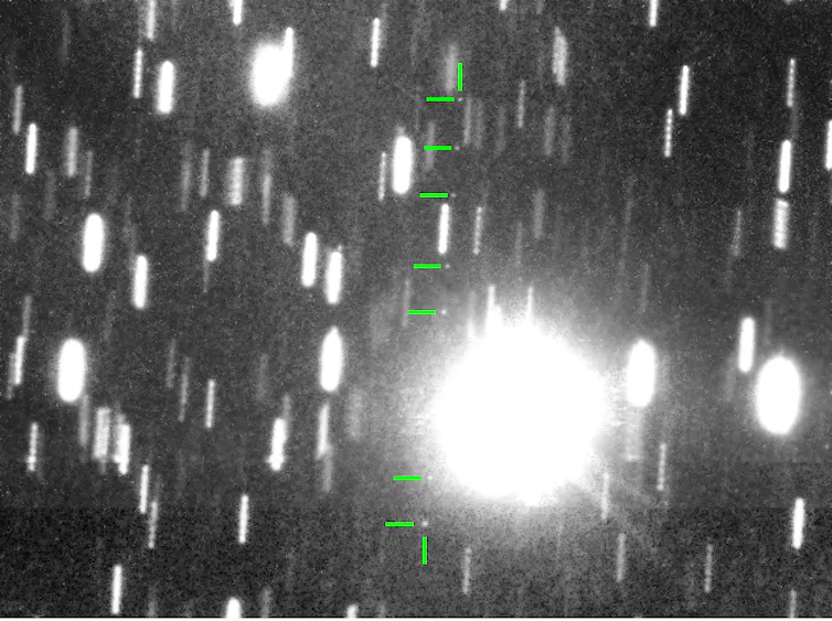 An image of the sky where stars appear as short streaks. One bright star appears as a large blob. Faint dots that track across the sky are marked with green arrows. These dots are the asteroid as it moves across the sky.