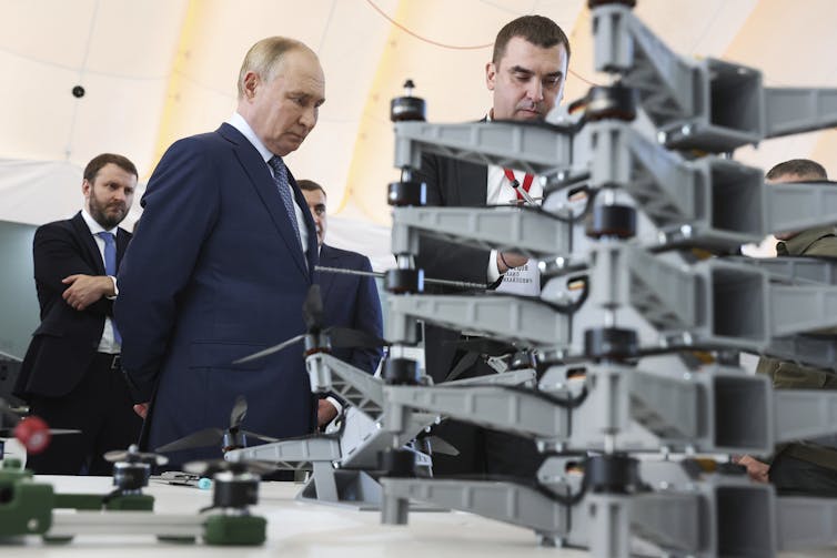 A balding man stares at a collection of drones as men stand around him.