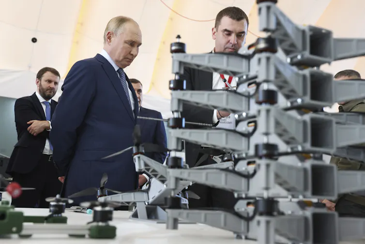 A balding man stares at a collection of drones as men stand around him.