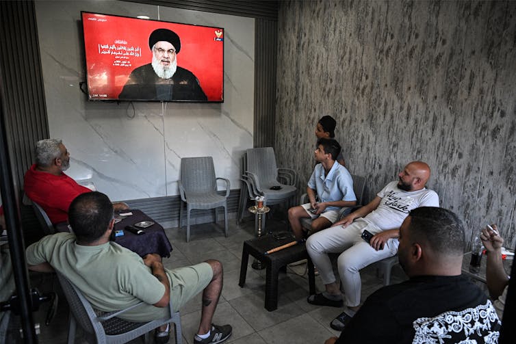 A group of people sit on chairs and watch a screen on which a man in a beard is talking.