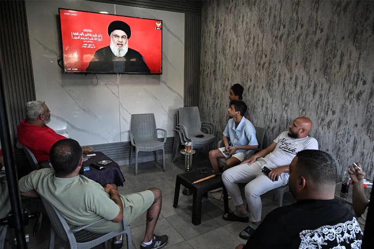 A group of people sit on chairs and watch a screen on which a man in a beard is talking.
