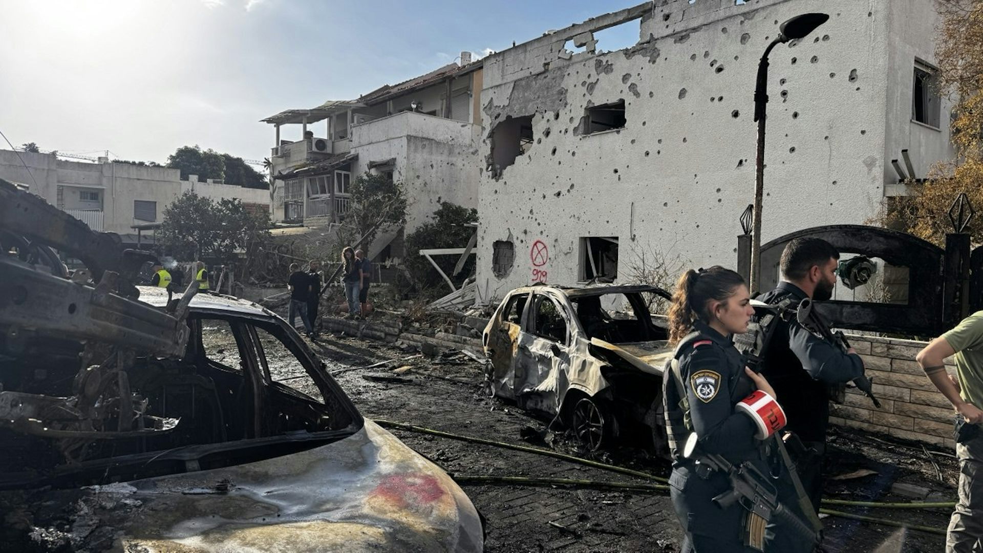 A burnt-out car can be seen in front of a damaged building facade.