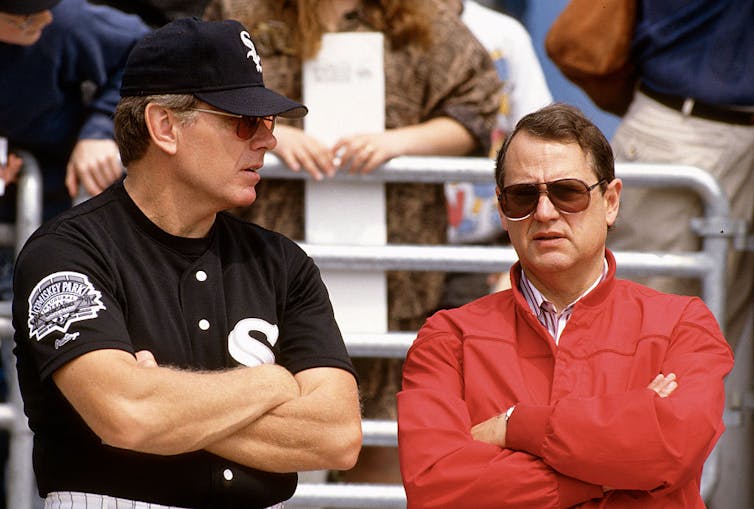 Man in black and white baseball uniform crossing his arms and chatting with man wearing sunglasses and a red jacket.