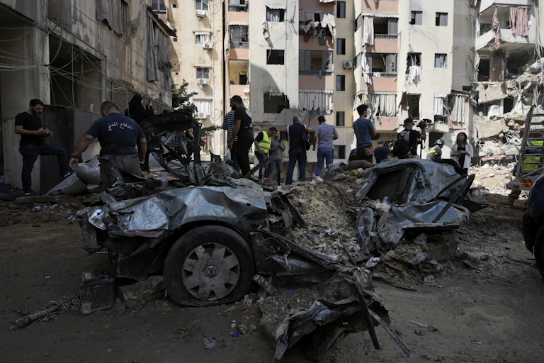 People stand near the wreckage of a blown up car. Damaged buildings are seen in the background