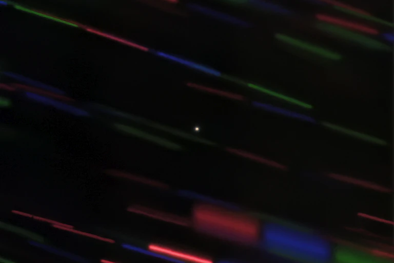 A black image with green, red and blue streaks (background stars). A small white dot (2020 CD3) is in the centre of the image.