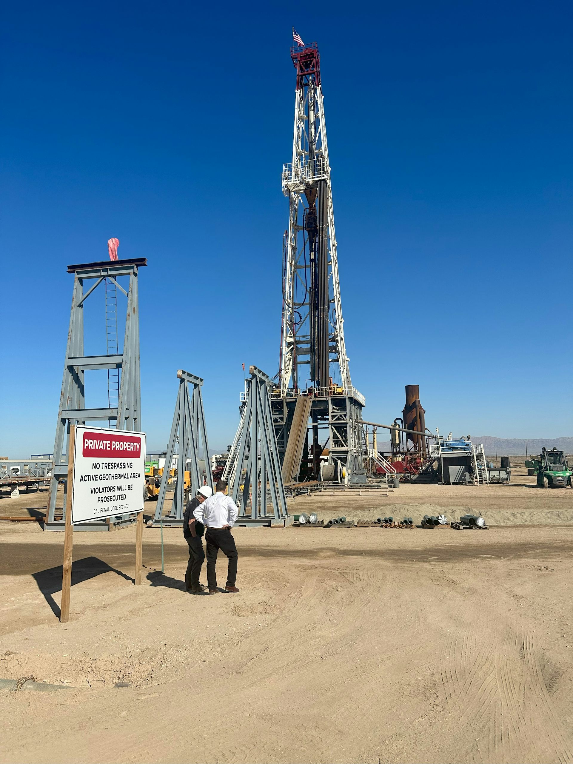 Two people stand near tall rigs used for pulling up briny water with lithium in it.