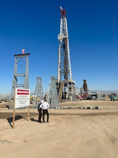 Two people stand near tall rigs used for pulling up briny water with lithium in it.