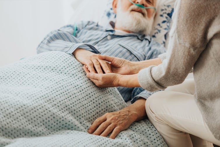 A senior man in bed. A woman holds his hand.