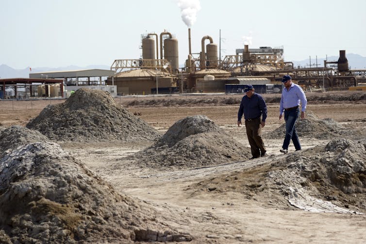 Two men walk among mud and sand mounds with an industrial facility in the background.