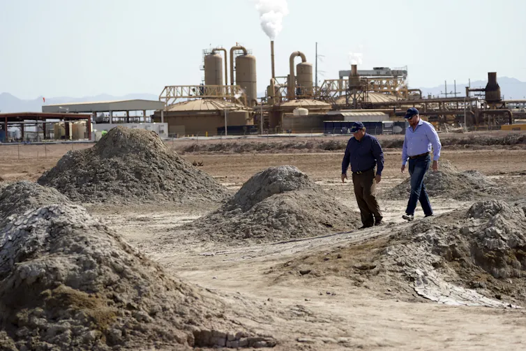 Two men walk among mud and sand mounds with an industrial facility in the background.