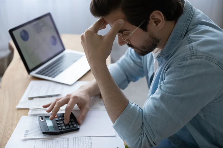 A young man holds his forehead as he looks at a calculator and bills.