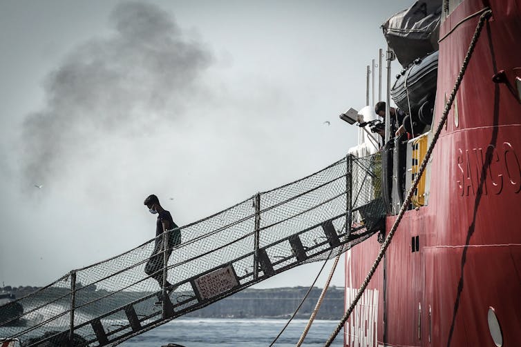 A man walks alone down the ramp off a red ship