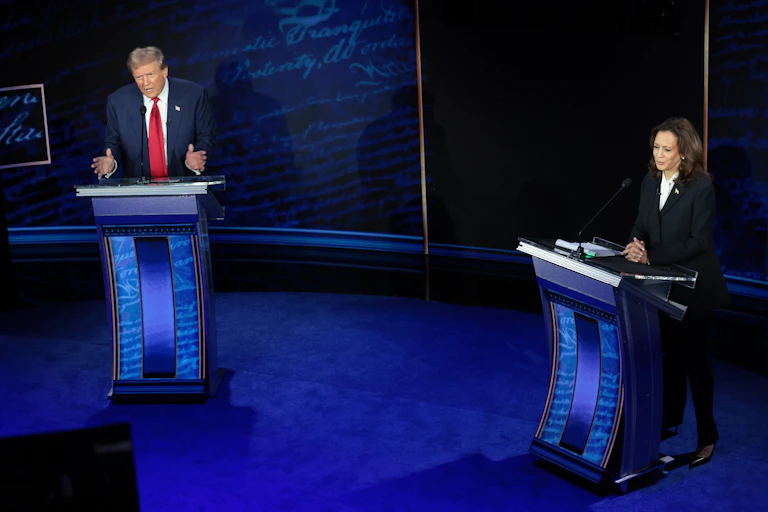 A man wearing a blue suit stands at a podium and gestures with his hands, nearby a woman in a dark suit who is also at a podium.