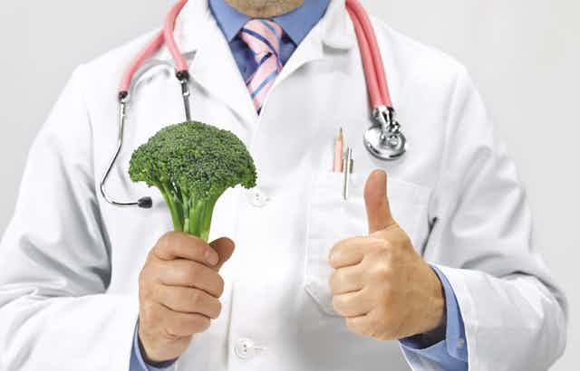 A doctor wearing a white jacket and a stethoscope around his neck is holding a stalk of broccoli in his right hand while giving a thumbs up with his left hand.