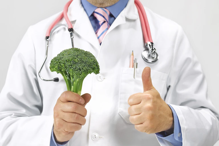 A doctor wearing a white jacket and a stethoscope around his neck is holding a stalk of broccoli in his right hand while giving a thumbs up with his left hand.