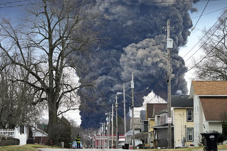 A huge plume of black smoke goes into a clear blue sky, with houses in the foreground and firefighter walking down the street.