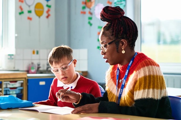 Sitting at a table in a classroom, a teacher instructs a student.