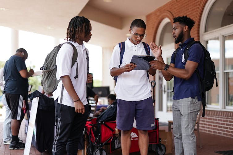 Three young men with backpacks, talking; one of them is reading something on a clipboard he's holding.