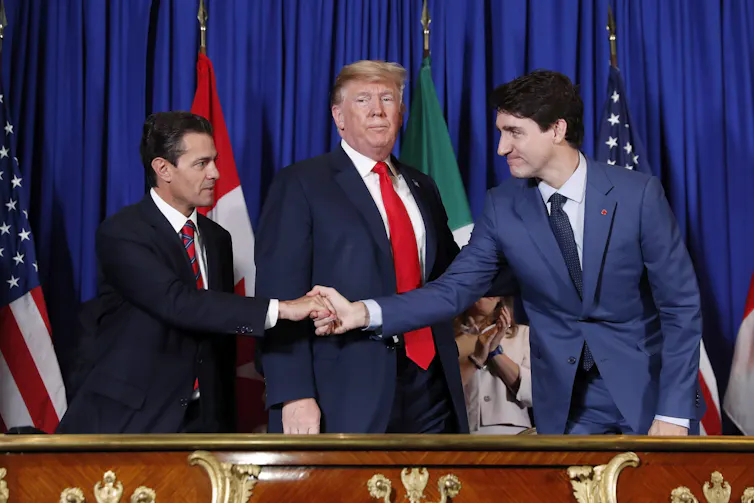 A rotund man with greyish-blond hair, wearing a blue suit and red tie, stands between two dark-haired men who are shaking hands in front of him.