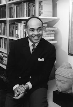 A Black man dressed in a business suit is smiling as he sits near shelves of books.