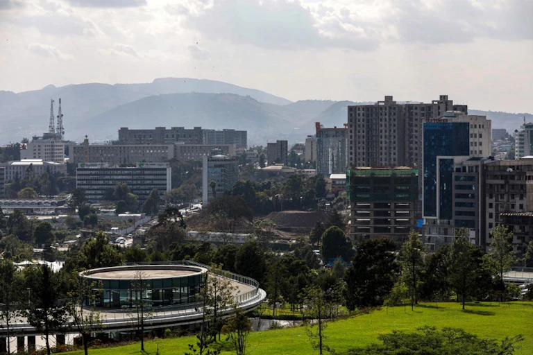 A manicured lawn in the foreground, high-rise buildings in the middle and hills in the background of a cityscape