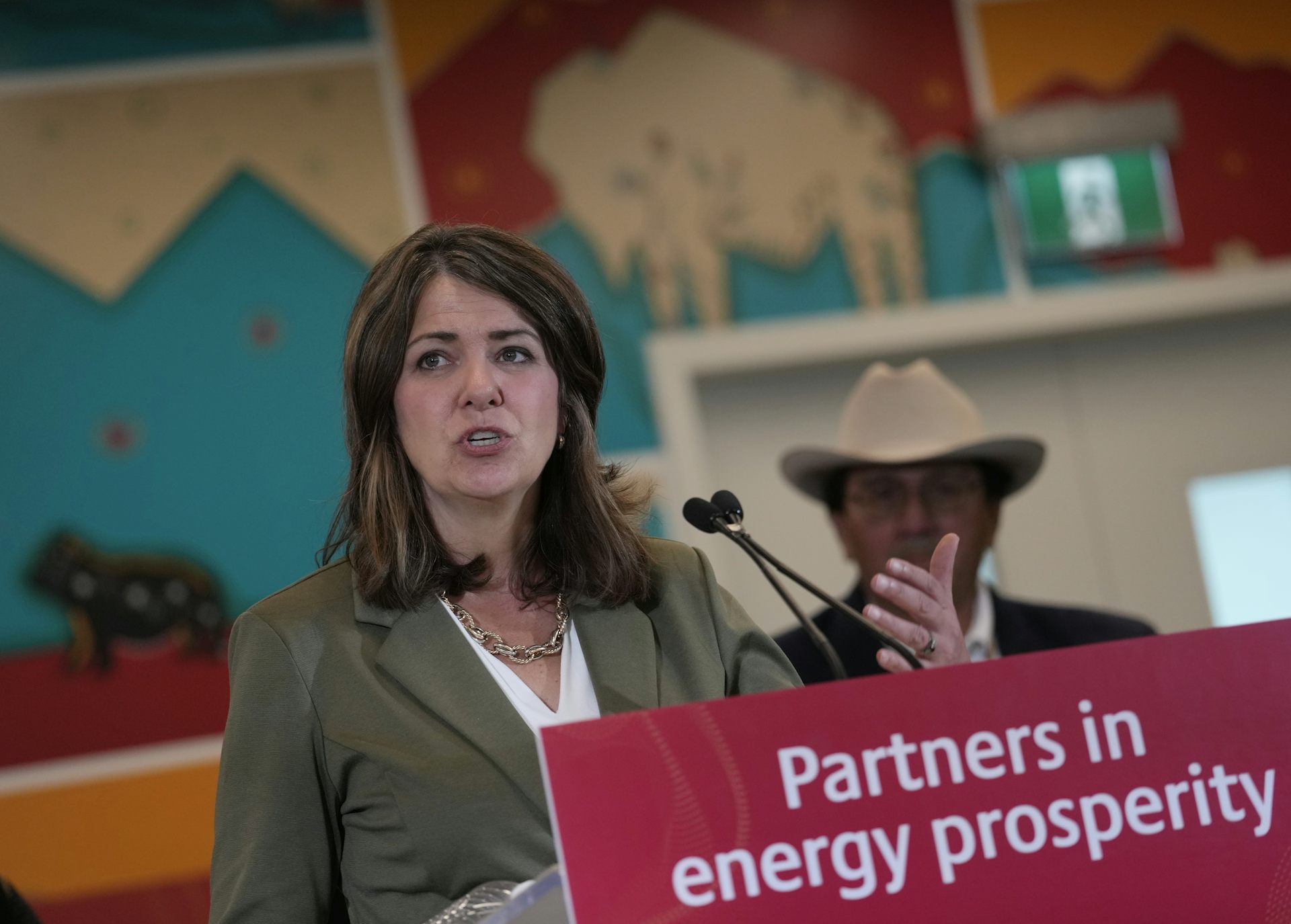 A woman with dark hair speaks into a microphone as a man in a cowboy hat stands behind her. A banner on the lectern in front of the woman reads 'partners in energy prosperity.'