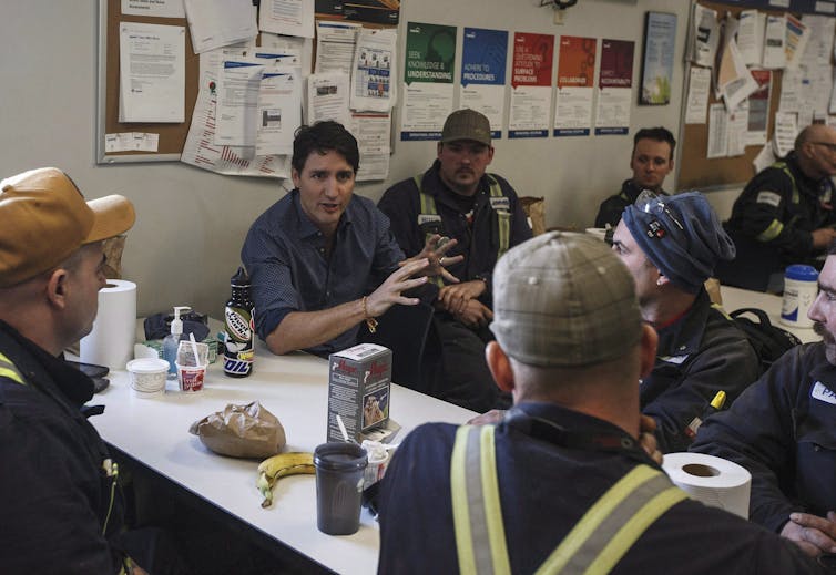 A dark-haired man sits at a table talking to workers.