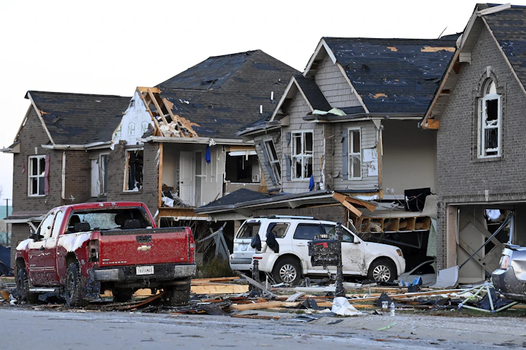 Trucks parked in front of four closely-built two-story homes, all with storm damage to roofs and windows.