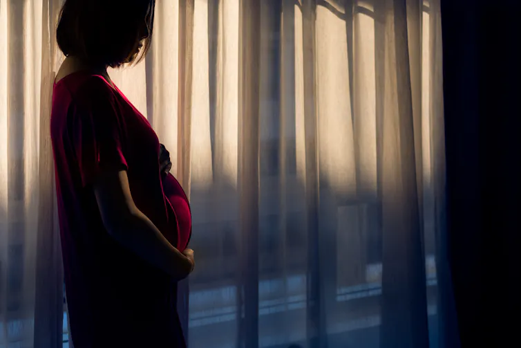 pregnant person stands in shadows hear window with curtain