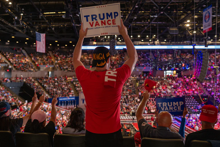 A man looks to a large crowd and wears a red shirt and holds a sign that says 'Trump Vance.' The stadium is filled with people and others hold signs that say 'Trump Vance.'