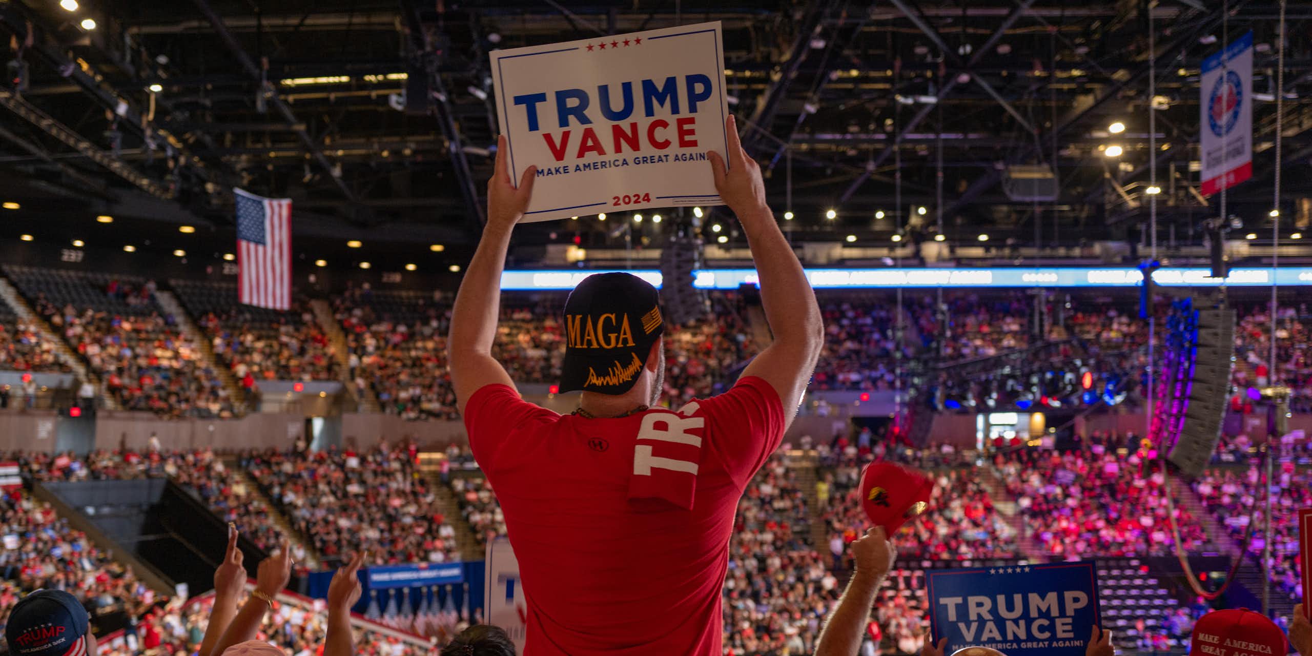 A man looks to a large crowd and wears a red shirt and holds a sign that says 'Trump Vance.' The stadium is filled with people and others hold signs that say 'Trump Vance.'