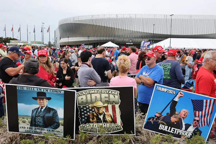 A crowd of people stand outside on a gray day, in front of three large signs that show a man dressed in a costume from the Wild West, with the words 'You tell em I'm coming,' and a poster of a man surrounded by men in suits as he raises his fist