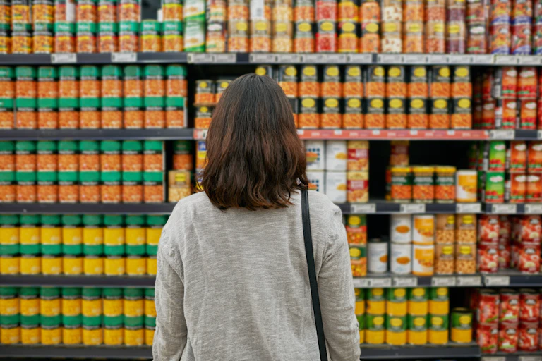 A woman stands in front of a row of canned goods in a supermarket