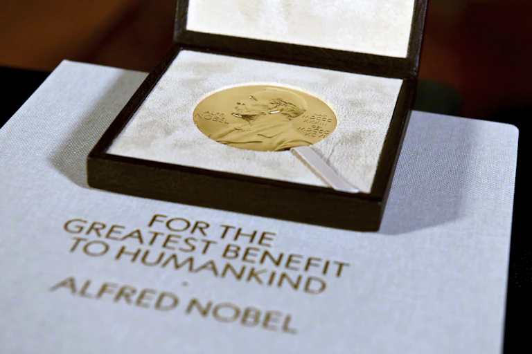 A nobel prize, which looks like a large coin, in a square case and a paper that says "For the greatest benefit to humankind, Alfred Nobel"
