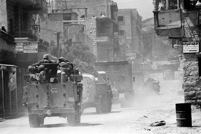Uniformed men in armored vehicles drive along a dusty road.