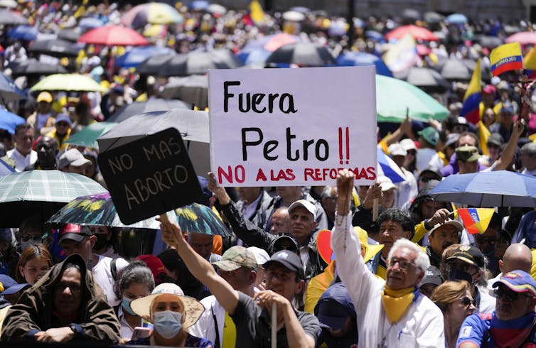 Protesters wave a banner reading Fuera Petro — fire Petro — at a protest on a sunny day in a city square.