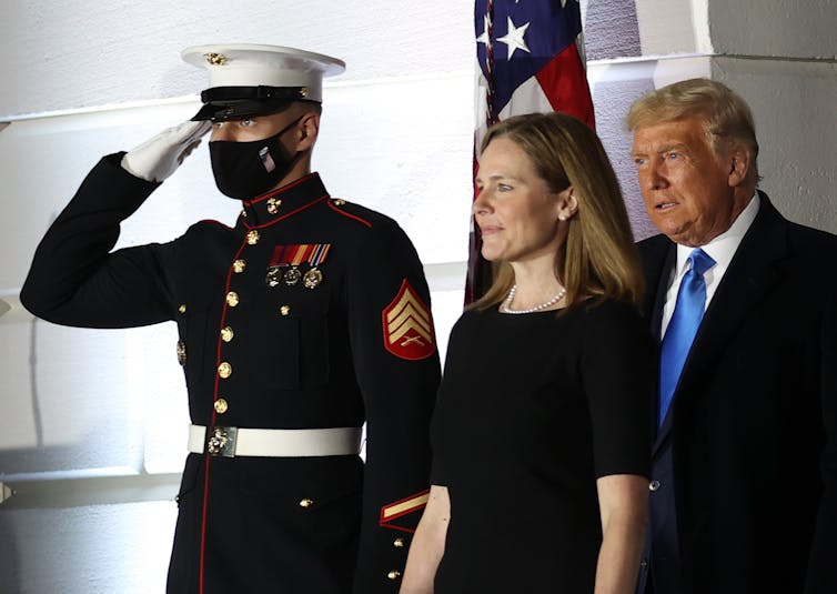 A man in military uniform saluting in front of a US flag as another man and woman walk past him.