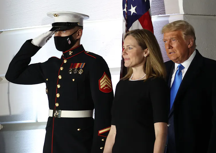 A man in military uniform saluting in front of a US flag as another man and woman walk past him.