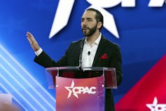 A dark-haired bearded man gestures as he speaks from behind a lectern.