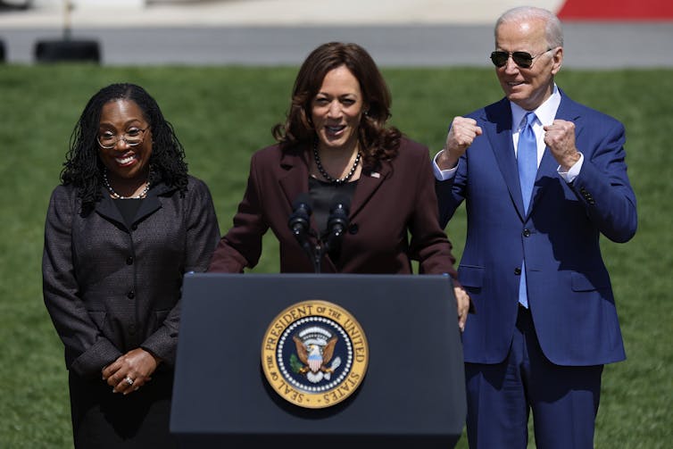 Two smiling women and one smiling man who is raising his fists in excitement, standing at a lectern outside.