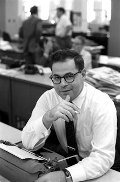 A dark-haired man with glasses wearing a white shirt and dark tie, sitting and smiling at a desk.