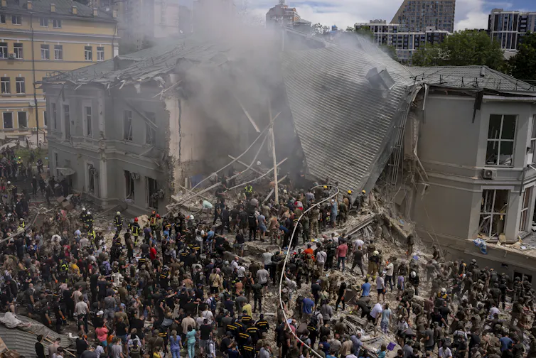 Crowds around a large building that has been bombed and partially destroyed.