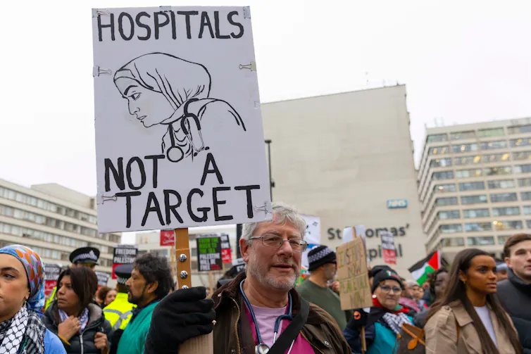 A man in a crowd holds a sign with a drawing of a female doctor reading: Hospitals Not a Target.