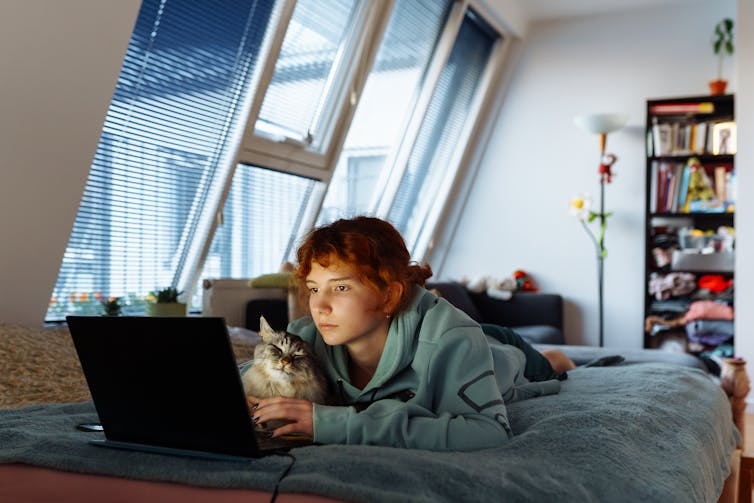 A teenage girl lies on a bed with a laptop and a cat.
