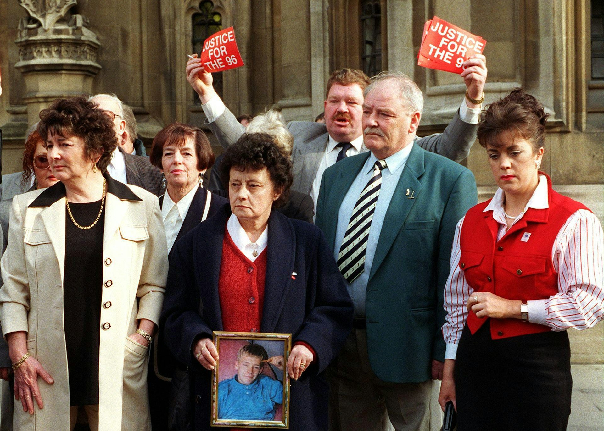A 1998 photo of a small group of Hillsborough family members before attending the Commons. A woman is holding a photo of her son, and one man is holding flyers that read 'justice for the 96'.