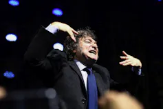 A man with shaggy brown hair grimaces while gesturing with his hands.