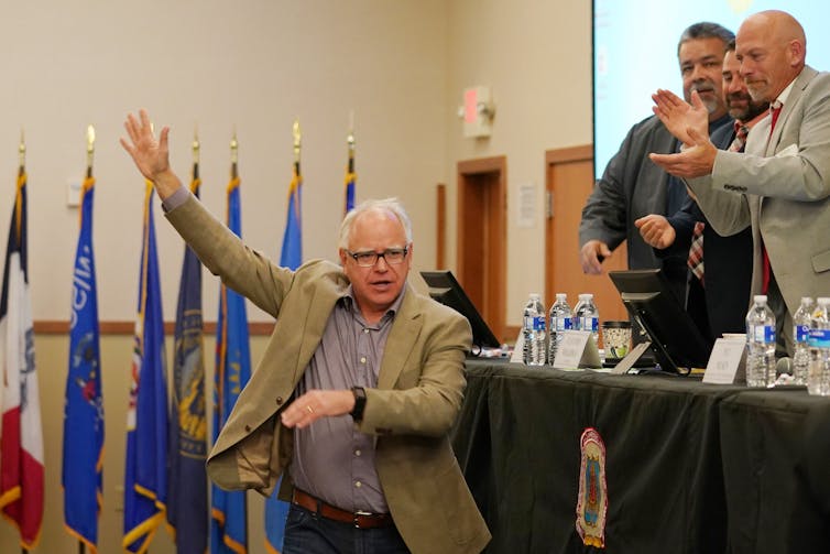 A white man with a brown blazer raises his arm and gestures while people nearby him clap. He stands in front of flags in an indoor room.